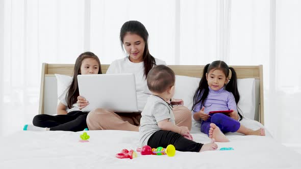 little daughter and Baby play baby toys together on bed while Busy mother working on laptop