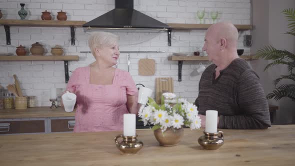 Happy Old Caucasian Lady Bringing White Cup with Tea or Coffee From Kitchen and Talking To Her alt