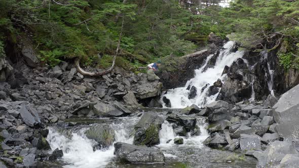 Backpacker Traveler Sits In Front Of Cascading Water Among Rocks Covered With Moss At Forest Park In alt