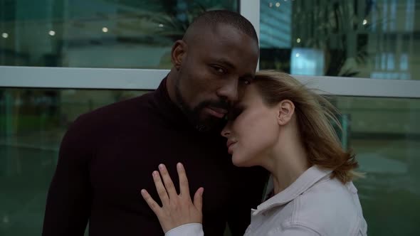 an African Man in Dark Clothing and a Blonde Woman in a Gray Jacket Are Hugging Each Other and alt
