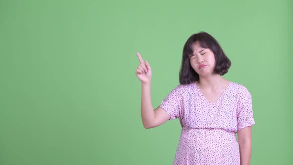 Stressed Asian Pregnant Woman Pointing Up and Giving Thumbs Down alt