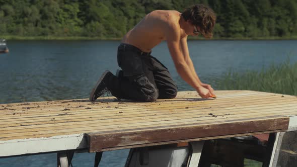 Young male carpenter using sharp scraper planing wooden roof planking of timber boat by hand. WIDE S alt