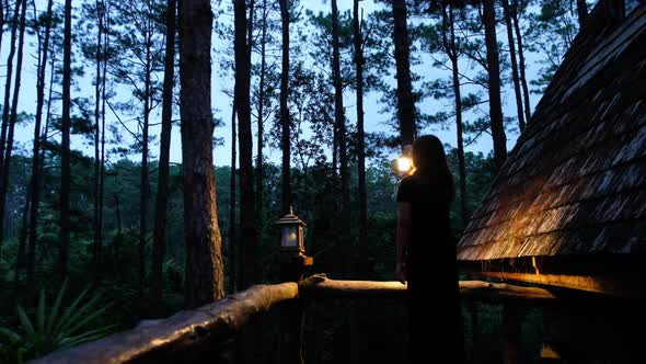 A young woman holding lantern and looking around the log cabin in the woods