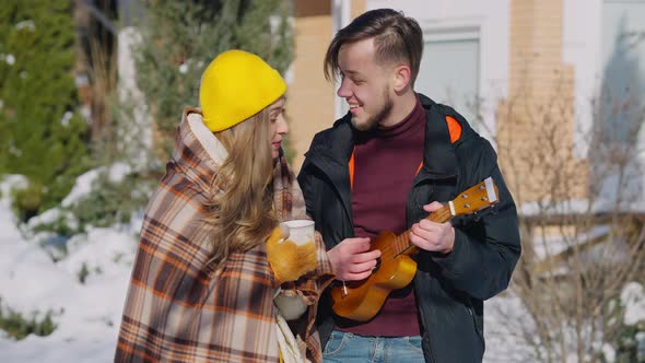 Young Man Playing Ukulele Singing Serenade for Smiling Woman Holding Coffee Tea Cup Outdoors on alt