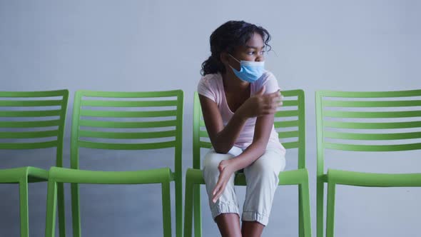 African american girl lowering her face mask while sitting on a chair at hospital alt