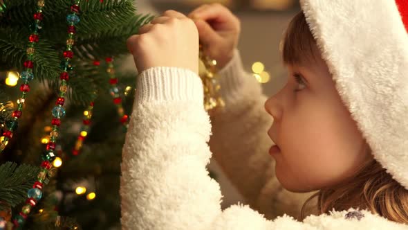 Little Girl in Red Santa Claus Hat Decorates Christmas Tree with Golden Bell