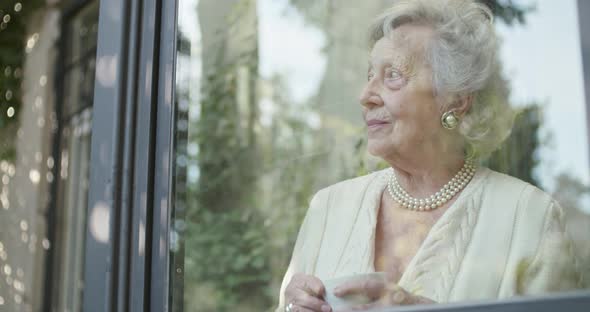 Senior Grandma Woman Enjoying Drinking Cup of Tea or Coffee Near Window Looking Outside