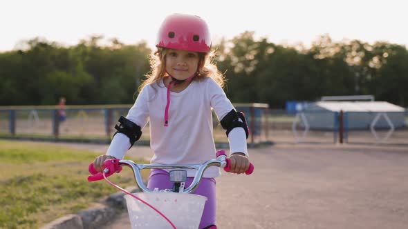 Cute Light Hair Little Girl in Pink Helmet in Elbow and Knee Pads Rides a Bicycle at the Street