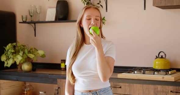 Young blonde woman is throwing up green apple and eating it in the kitchen. alt