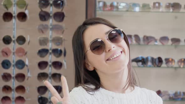 Cheerful Young Woman in Sunglasses Smiling on the Background of a Shelf with Glasses in the Store alt