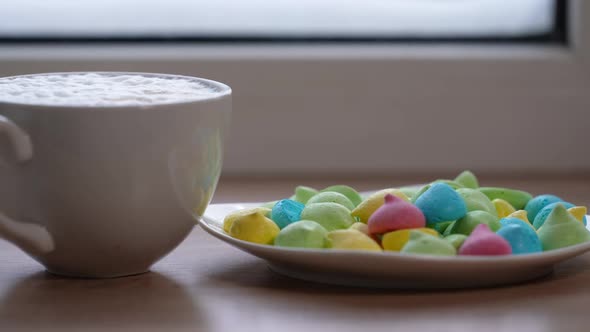 A Large Mug of Coffee with Milk or Mochacino and Colorful Sweets in a Plate on the Windowsill in