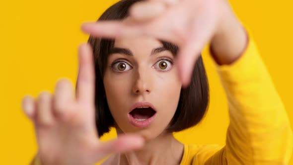 Young Woman Looking At Camera Through Finger Frame Yellow Background alt
