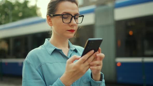 Woman Stands at a Public Transport Stop and Using Smartphone alt