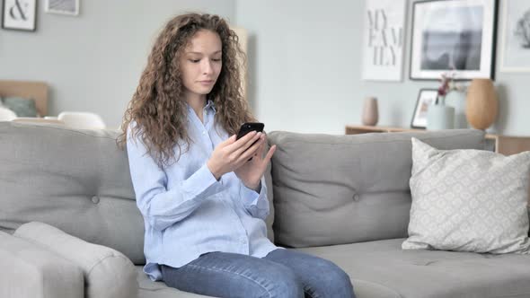 Young Curly Hair Woman Using Smartphone While Relaxing On Sofa By