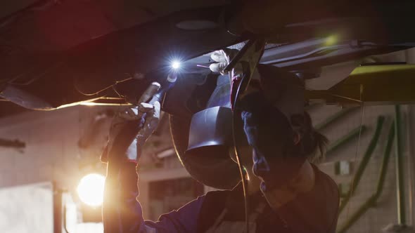 Female mechanic wearing welding helmet welding under a car at a car service station alt