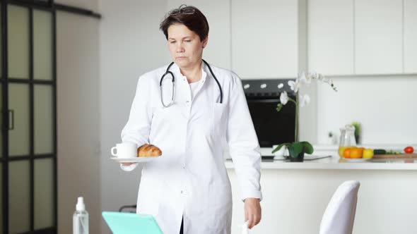 Portrait of Tired Female Doctor Walking to Table with Tablet Holding Tea and Croissant alt
