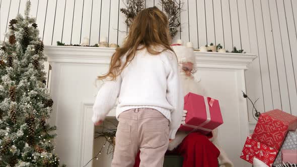 Little Girl Bringing a Present for Santa and Hugging Him in His Christmas Workshop alt