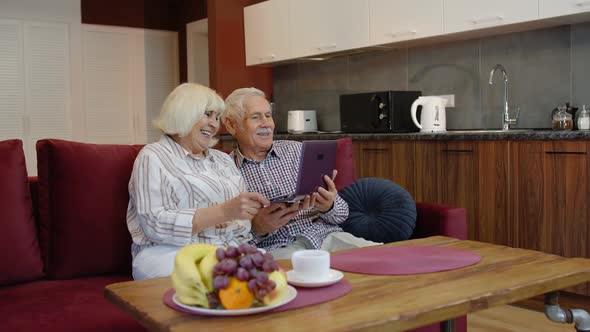 Senior Pensioner Couple with Digital Laptop Pc Computer at Home. Resting on Sofa in Cozy Living Room alt