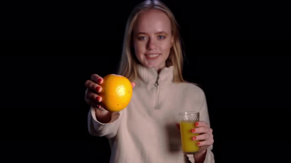 Young Pretty Caucasian Blond Woman Holding a Glass with Orange Juice and Orange Over Black