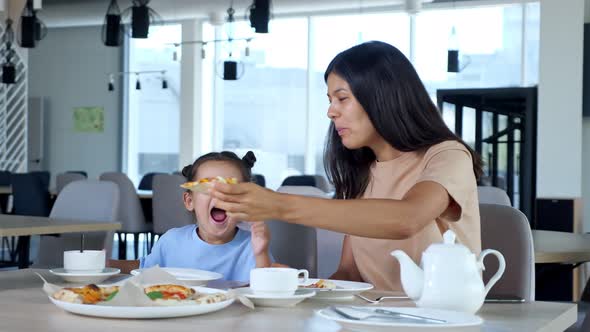Young Mom Holds Pizza Slice and Child Girl Eats Playing alt