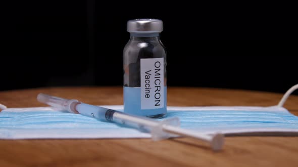 Ampoule with Omicron vaccine, syringe and medical mask on wooden background alt