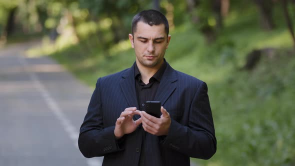 Serious Adult Caucasian Man User Stand Outdoors in Park Wears Black Formal Suit Hold Mobile alt