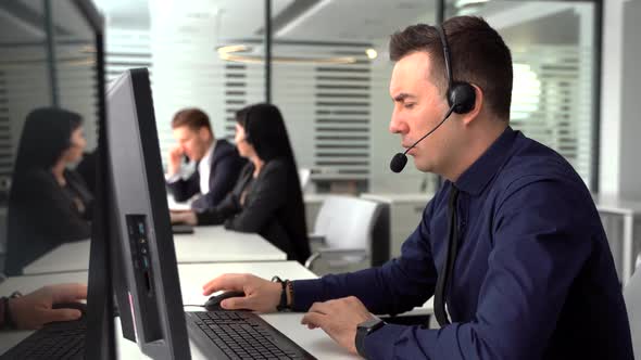 Smiling Man Taking Call in Busy Call Center. In the Background There Are His Colleagues Also Talking