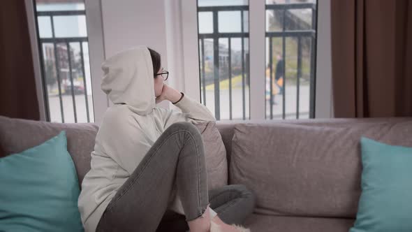 Girl in the Hood Looks Thoughtfully Out the Window While Sitting on the Sofa alt
