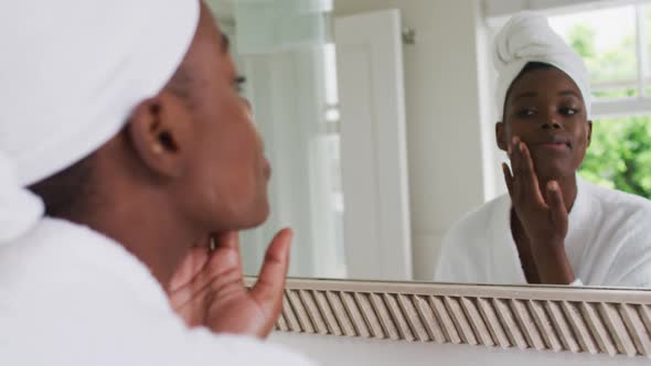 African american woman in bathrobe applying face cream while looking in the mirror at home alt