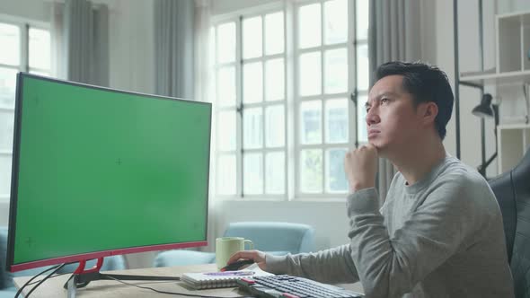 Asian Man Thinking Then Typing On Green Screen Desktop Computer While Working At Home alt