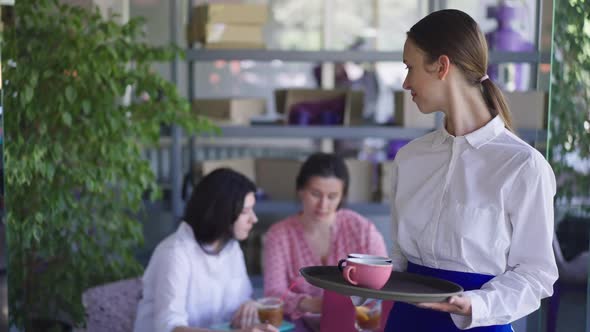 Young Slim Waitress with Coffee Cups on Tray Looking Back at Clients Sitting at Table Turning to alt