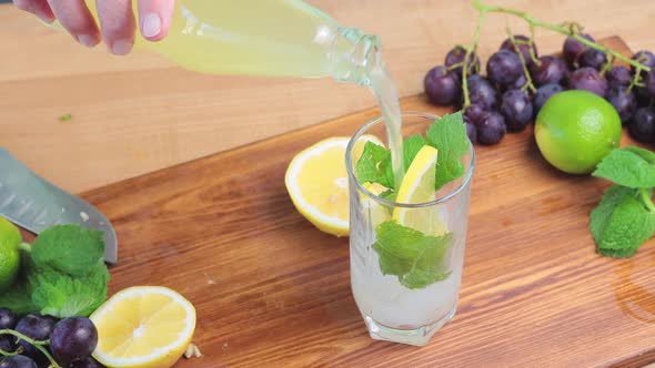 Pouring lemonade from bottle into glass with mint, ice cubes and lemon slices, close up view