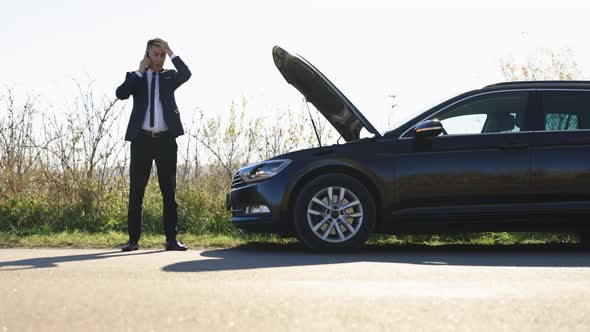 Mature Business Man in Black Suit Standing Near Broken Car with Open Hood and Talking on Mobile