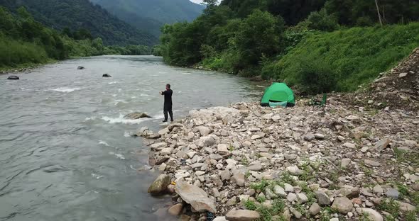 A Fisherman Fishes By Spinning a View of a Mountain River and a Green Tent alt