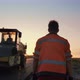 A Male Road Worker in Uniform Walks to a Road Roller in the Rays of the Setting Sun - VideoHive Item for Sale