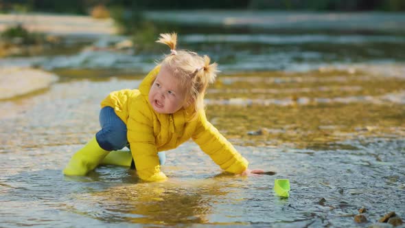 Little girl in yellow rubber boots and jacket standing in the water and plays with paper ship alt