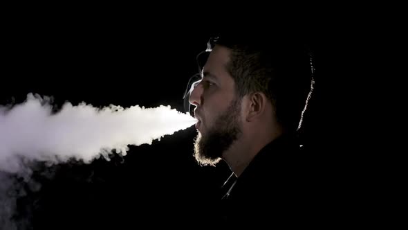 A young white bearded man in black shirt is vaping an electronic cigarette. alt