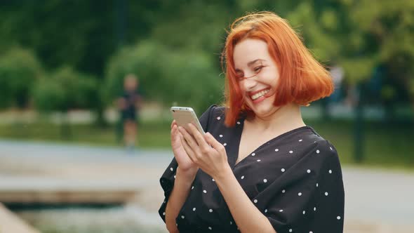 Attractive Redhead Caucasian Young Girl Woman Stands in Summer Park Looks in Mobile Phone Device alt