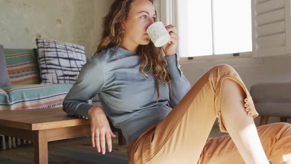 Thoughtful caucasian woman sitting on floor in living room drinking coffee, looking out of window alt