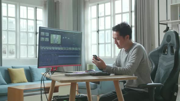 Asian Video Editor Man Using Mobile Phone While Using Desktop Computer For Editing Video At Home alt
