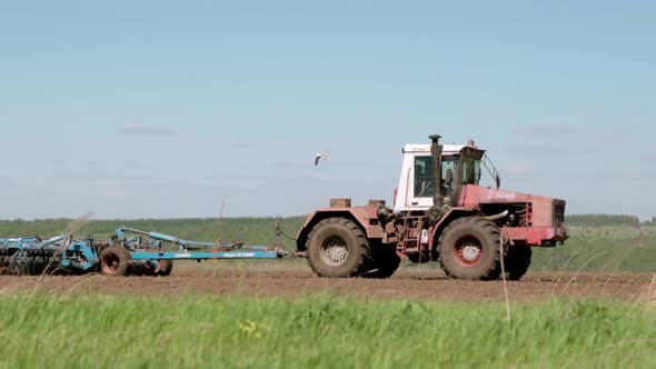The Tractor Cultivates the Land in the Field Against the Backdrop of a Beautiful Green Forest alt