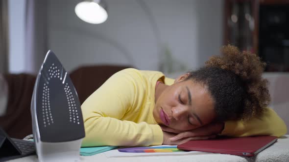 Exhausted Young African American Woman Sleeping on Laptop on Ironing Board in Home Office alt