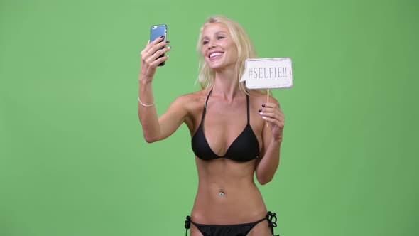 Happy Young Beautiful Blonde Woman Wearing Bikini While Taking Selfie with Paper Sign