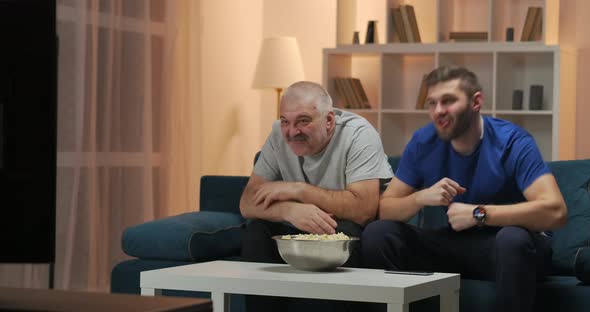 Father and Son Watching a Sports Match on TV While Sitting on the Couch in Their Home alt