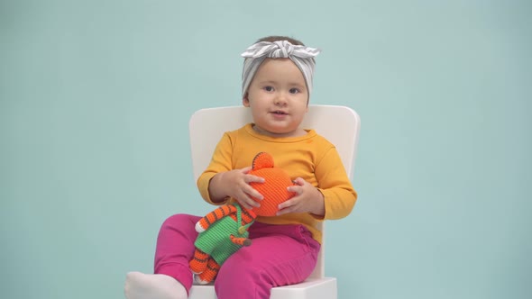 Toddler 12-17 months old sits on a small white chair and plays with a knitted toy tiger alt