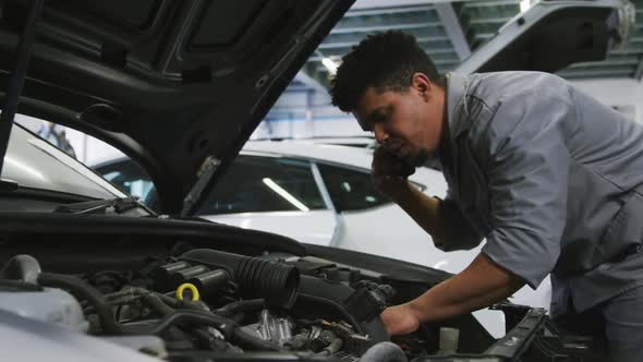 African American male car mechanic looking at an open car engine and talking on a smartphone alt