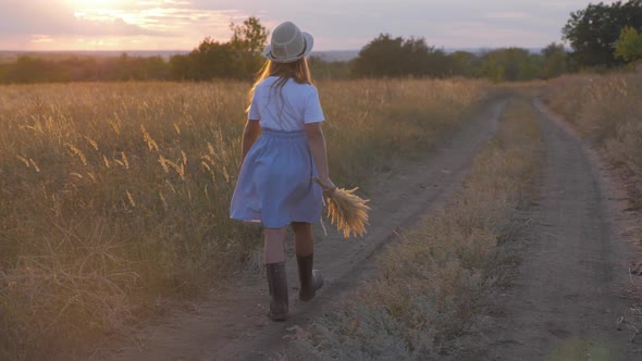 A Girl Walks Along a Rural Road at Sunset, Child in a Hat and Rubber Boots alt