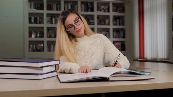A Student Girl with Glasses is Sitting at a Library and Writing in a Notebook alt