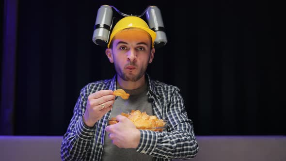 Young Funny Man With Beer Helmet on the Head Eating Chips While Watching Movie on the Sofa