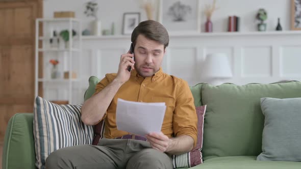Young Man Angry On Smartphone while Reading Papers on Sofa alt